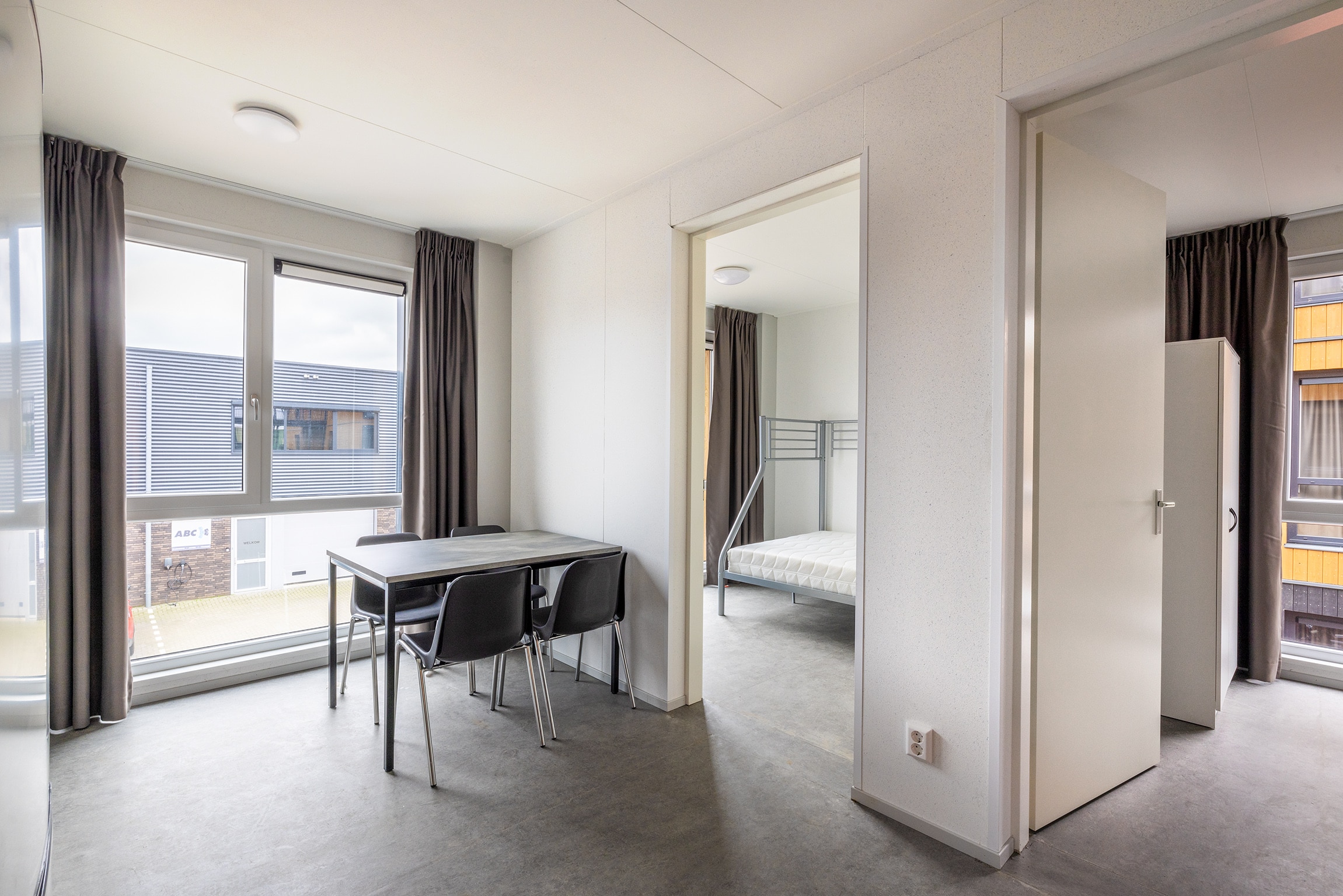 Binnenaangezicht met tafels en stoelen en slaapkamer met stapelbed op de achtergrond.